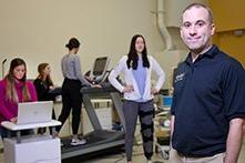 A man smiling at the camera while a woman walks on a treadmill and others perform fitness evaluations.